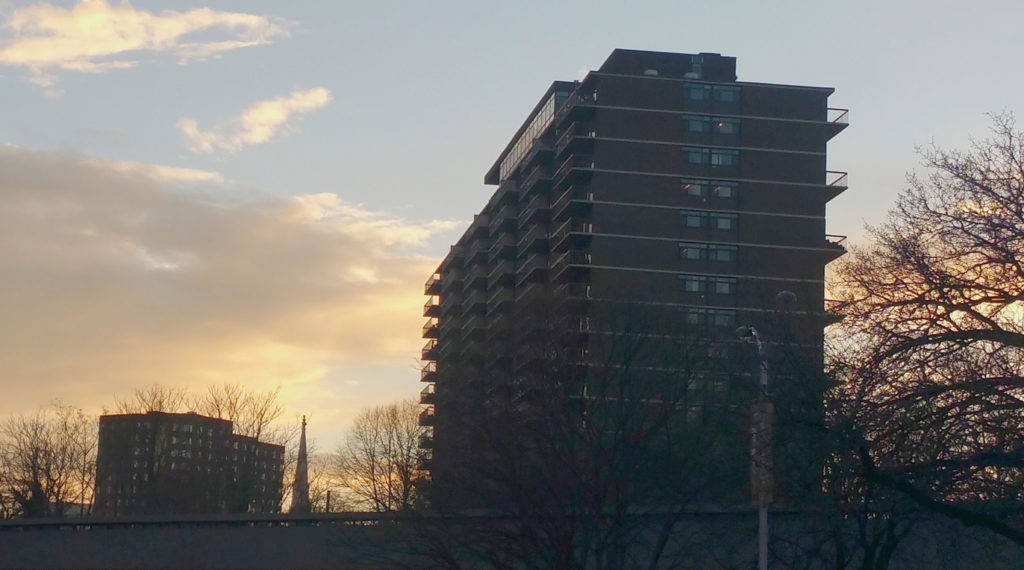 Sunset view of Bolton Hill (Baltimore, MD) featuring blue sky, white clouds and bare trees. High-rise, residential building and overpass seen in foreground; high-rise, residential building and church steeple in background.