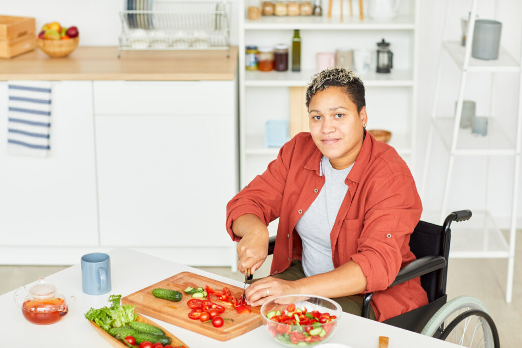 Portrait of disabled young woman with short hair looking at camera while sitting in wheelchair and cutting vegetables in the kitchen